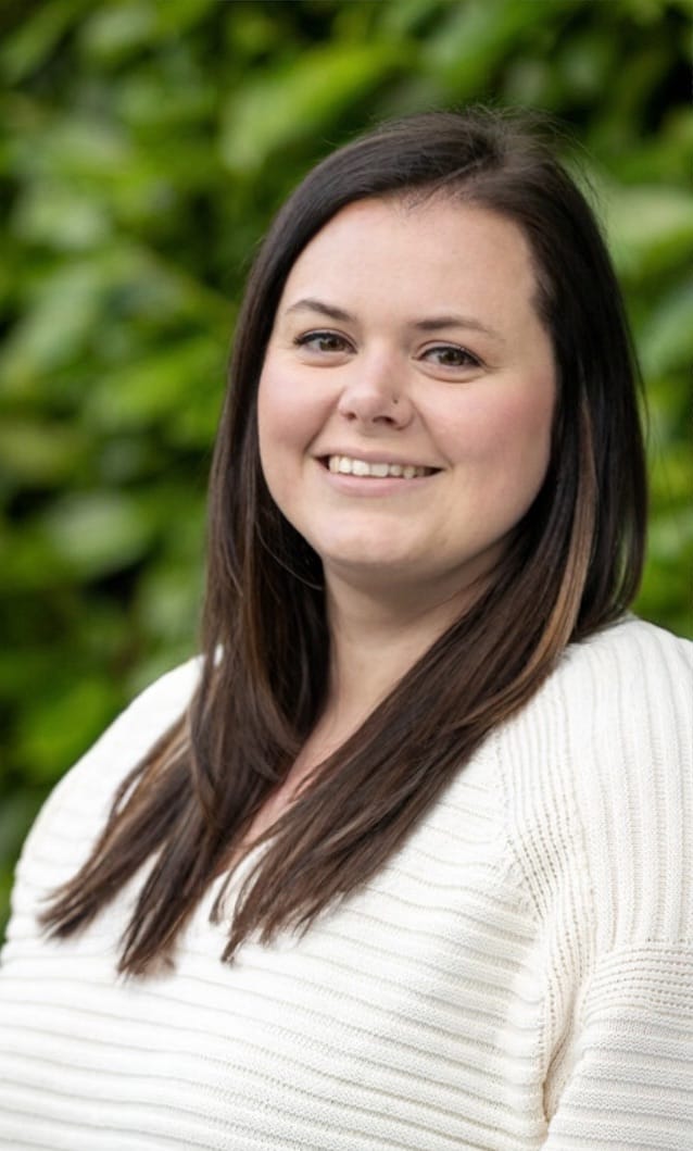 A woman with long, dark brown hair, in a green top, stood outside next to a tree.